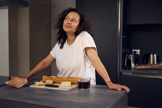 Multi ethnic pensive happy woman smiling looking away, standing at kitchen counter with a cup of freshly brewed coffee and fresh sandwiches for breakfast