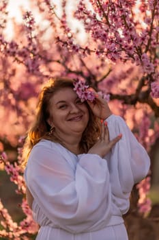 Woman blooming peach orchard. Against the backdrop of a picturesque peach orchard, a woman in a long white dress enjoys a peaceful walk in the park, surrounded by the beauty of nature