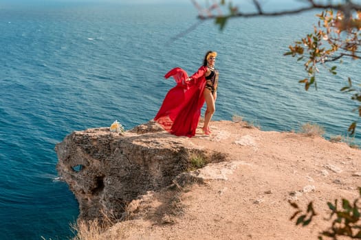 A woman in a red dress is standing on a rocky cliff overlooking the ocean. She is wearing a lei and she is dancing or celebrating. The scene is serene and peaceful, with the ocean as a backdrop