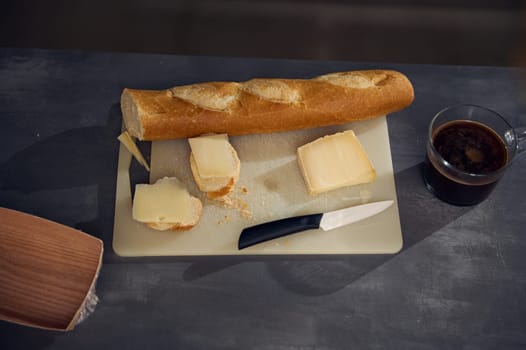 Still life top view breakfast on the kitchen table. A cup of freshly brewed espresso coffee near a cutting board with a whole grain baguette, loaf of bread and sliced cheese on the kitchen table