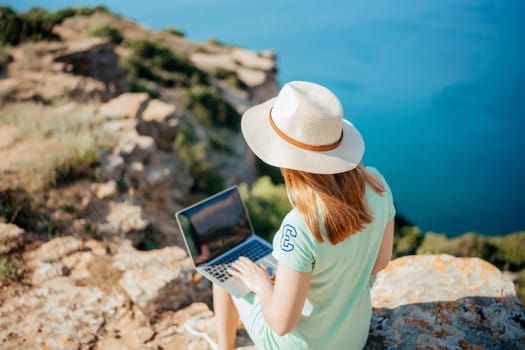 A woman is sitting on a rock with a laptop open in front of her. She is wearing a straw hat and a green shirt. The scene suggests a relaxed and leisurely atmosphere