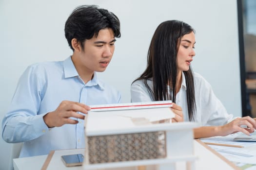 Professional male asian architect using ruler to measure house model length while young beautiful caucasian colleague using laptop to analyzed data on meeting table with house model. Immaculate.