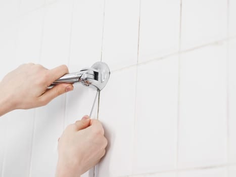 The hands of a young caucasian guy unscrew the screw with a small hex wrench to remove an old metal hanger with long tubes on a white tiled wall in the bathroom, close-up view from below. The concept of dismantling, repair, cleaning services, at home, bathroom, replace the hanger.