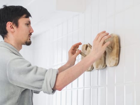 A young handsome caucasian guy in a gray fleece bathrobe with rolled up sleeves hangs a new wooden stylish and eco-friendly hanger on a wall with white tiles in the bathroom, close-up from below. The concept of installing a hanger, installation, dismantling, repair, cleaning the house, bathroom, eco-friendly accessory.