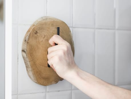 One young unrecognizable Caucasian guy installs a wooden round cut towel rack with one hand on a white tiled wall in the bathroom on a summer day, side view close-up.