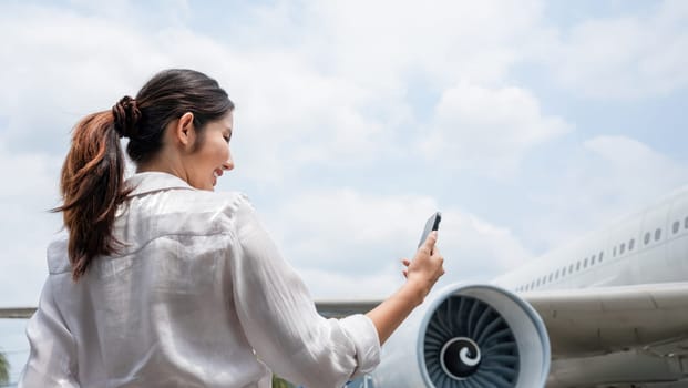 Asian woman smiling while using smartphone near airplane at the airport. Concept of travel, technology, and modern transportation..