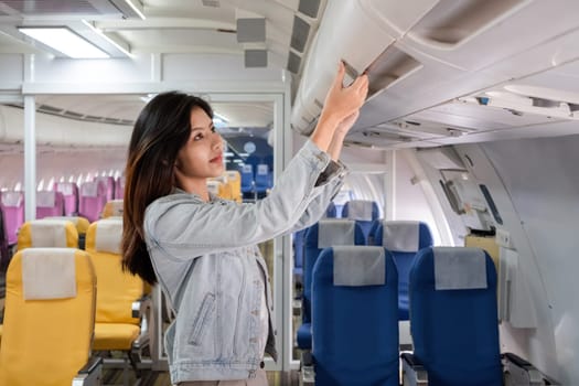 Asian woman adjusting overhead bin in airplane cabin. Concept of travel, aviation, and in-flight preparation.