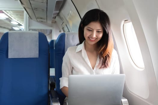 Asian woman using laptop on airplane. Concept of travel, technology, and productivity.