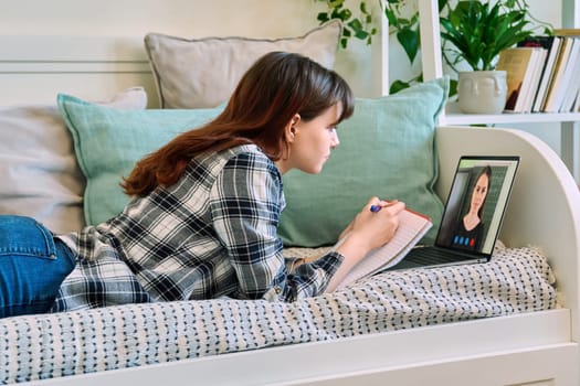 Female student lying on couch at home, looking at laptop computer screen, having remote video lesson, consultation, listening to online course, writing in notebook. Technology education study youth