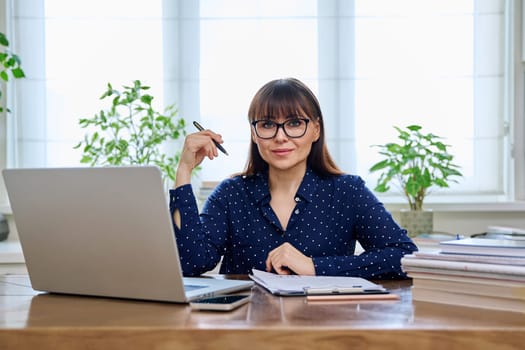 Middle-aged smiling woman working with laptop computer business papers, sitting at desk in home office interior, looking at camera. Work, remote business, freelance, technology, 40s people concept