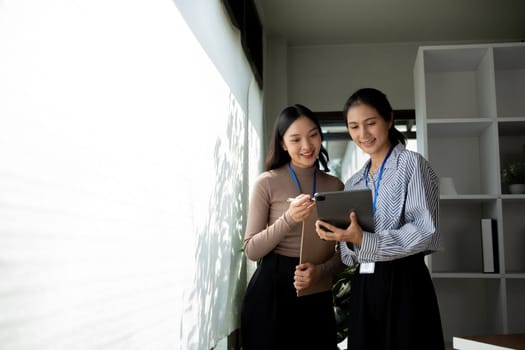 Two Asian businesswomen discussing work using a tablet in a modern office. Concept of teamwork and professional collaboration.