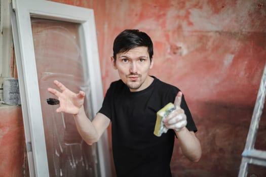 Handsome caucasian young brunette man emotionally speaks to the camera waving his hands with a bandaged finger with a black adhesive tape and a soapy sponge against the background of a window frame and a red wall, close-up side view with selective focus. The concept of home renovation, washing window frames.