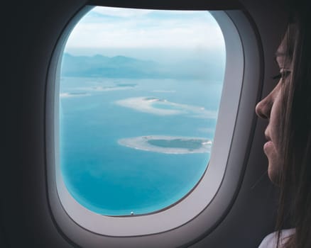 A woman looks thoughtfully out of an airplane window, observing pristine islands surrounded by blue waters below, as she travels during the middle of the day.