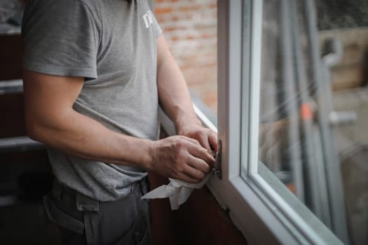 Caucasian man in gray uniform cleans his hands with dry paper napkin inside window frame opening, close up side view with selective focus. The concept of home renovation, washing window frames.
