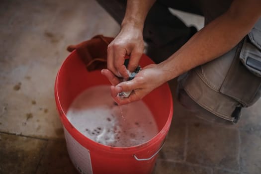 Hands of a caucasian man washes with a sponge with metal soap detail from a window frame over a red bucket of water while squatting in a room where repairs are underway, top view close-up with selective focus.