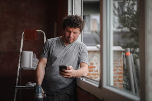 One young handsome caucasian brunette man holds a mobile phone and looks at the screen with a smile while standing at the window, side view close-up.