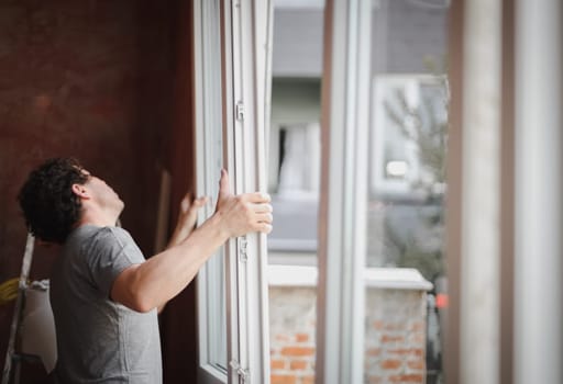 Young handsome caucasian man with curly hair installs a window frame with glass in a room where renovations are underway, close-up side view.