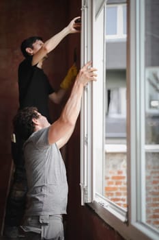 Two young handsome caucasian brunette men installs a window frame with glass on hinges while standing sideways in a room where repairs are underway, side view close-up.