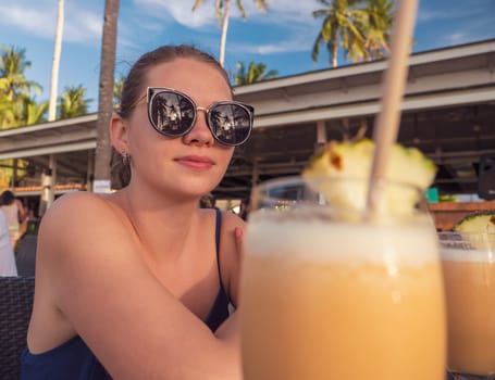 A woman wearing sunglasses is sitting at a beachside cafe, relaxing with a couple of pina coladas as the sun sets. The scene features palm trees and a laid-back atmosphere, perfect for unwinding.