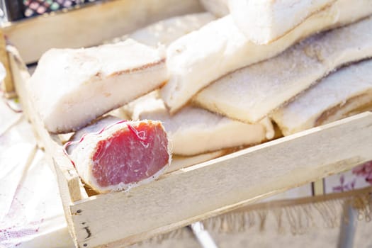 Various homemade pork products, including lard, balyk, and polendvitsa, are displayed on a rustic wooden tray at an outdoor market. The products are showcased in natural light, highlighting their freshness and quality.