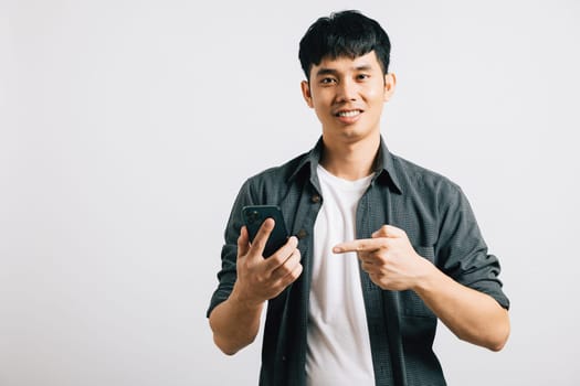 Smiling young man makes a pointing gesture at his smartphone screen for online shopping. Studio portrait isolated on white, showcasing confident technology use. Copy space is featured.