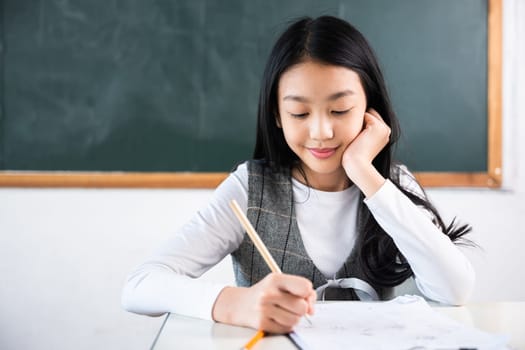 A girl is sitting at a desk and writing with a pencil. She is smiling and she is enjoying herself