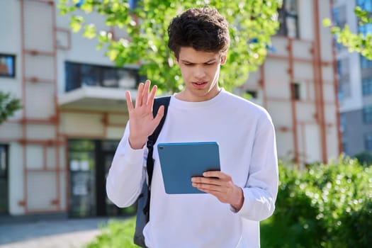 Young handsome guy college student using digital tablet outdoor, talking on video call chat conference, educational building background. Education, technology, training, 19,20 years age youth concept