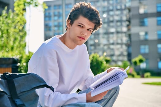 Handsome young male university college student with book looking at camera sitting on outdoor steps. Education, knowledge, youth 19-20 years old, literary hobby and leisure