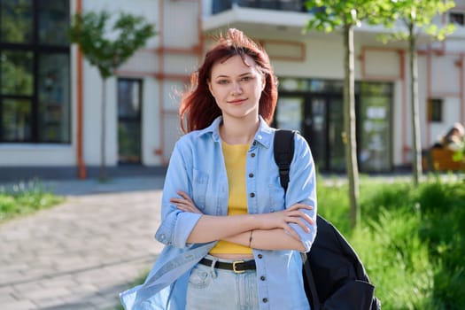 Portrait of young beautiful female college student outdoor, smiling red-haired confident girl with crossed arms backpack looking at camera, educational building background. Education training youth