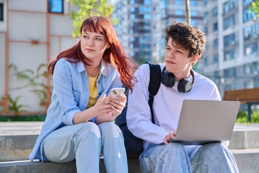 Teenage college students guy and girl talking, using laptop smartphone, sitting outdoor near educational building. Youth 19-20 years old, education, technologies, lifestyle, friendship concept