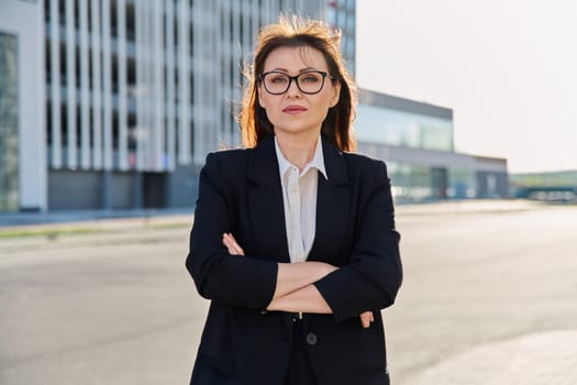Portrait of middle-aged business confident woman in suit, with crossed arms, posing outdoors in urban style. Office employee worker businesswoman teacher entrepreneur economist banker financier lawyer