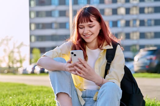 Young beautiful red-haired woman using smartphone, sitting on floor, sunny day outdoor city urban style. Youth, lifestyle, technology, internet online applications for leisure tourism learning work