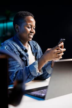 African American female student engaged in late night study session sits at desk, laptop open, and chats with group of fellow students on her mobile device. Close-up of joyful woman using cellphone.