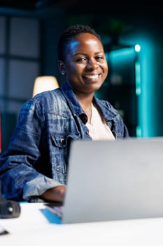 Portrait of African American blogger works on a laptop in a modern apartment. Smiling black woman is a dedicated freelancer, typing and reviewing information for her project.