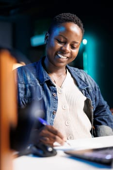 Close-up of african american female blogger jotting down notes, thinking of new ideas for online content. Happy black woman sitting at desk, using pen and paper for studying and reviewing project.