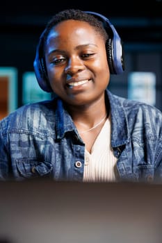 Close-up of african american lady wearing headphones while looking at her digital laptop, watching movies. Portrait of smiling black woman with a wireless headset gazing at her device screen.