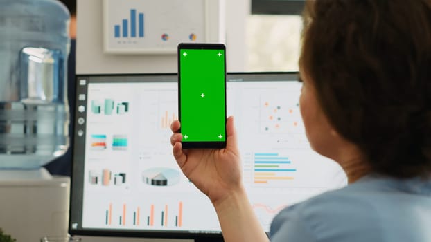Businesswoman looks at greenscreen on phone while she checks marketing renevue in coworking space, employee holding mobile phone display showing isolated copyspace template. Specialist at job.