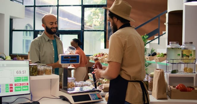 Merchant at checkout scanning and weighting goods to sell to frequent customer in local zero waste eco store. Young adult putting vegetables on scale, selling locally grown produce.