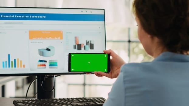 Business specialist shows greenscreen on smartphone software display, looking at isolated chromakey template and working on computer at desk. Using blank mockup and finishing agency operations.