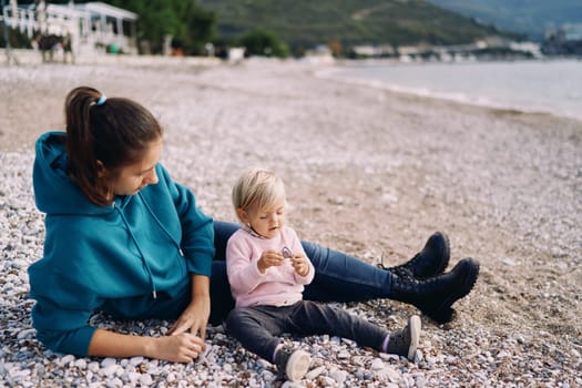 Mom lying down watching a little girl playing with pebbles sitting on the beach. High quality photo