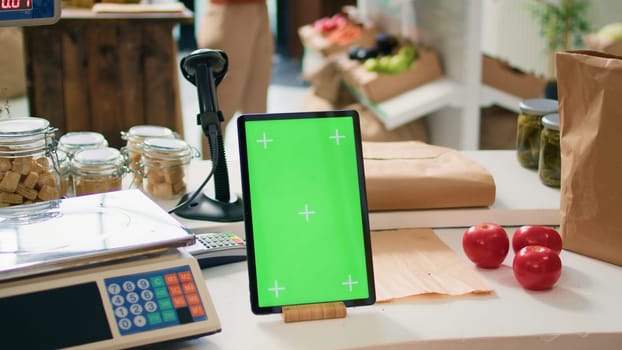 Greenscreen on tablet at checkout desk in local organic shop with freshly harvested produce, vendor using isolated chromakey template on gadget. Shop owner shows blank mockup display.