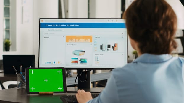 Office worker checks greenscreen display on tablet, working on business operations with isolated copyspace template and analyzing data on computer at desk. Having gadget with mockup.