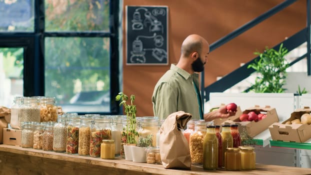 Client entering local grocery store to buy organic fruits and vegetables, looking at pasta and grains stored in sustainable nonpolluting glass jars. Middle eastern man advertising healthy eating.