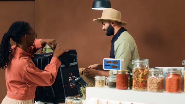 Merchant gives food order for delivery, putting homegrown organic vegetables in backpack. African american courier coming to pick up bio supermarket merchandise for local customers.