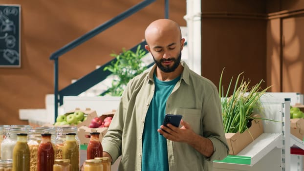 Middle eastern man checks ingredients list for pasta stored in nonpolluting glass containers, reading organic products benefits on smartphone. Vegan client ensuring about eco natural groceries.