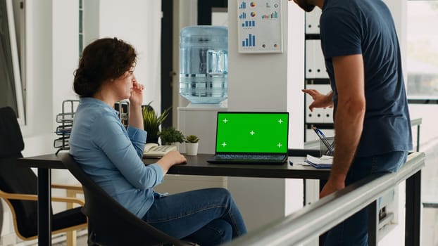 Colleagues looking at laptop on desk, analyzing isolated greenscreen layout in coworking space. Man and woman working in team at business office, using blank mockup chromakey template.