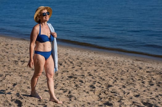 An old woman in a straw hat, sunglasses and a swimsuit is resting on the beach