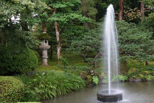 A serene Japanese garden featuring a stone lantern, lush greenery, and a tall water fountain in a pond.