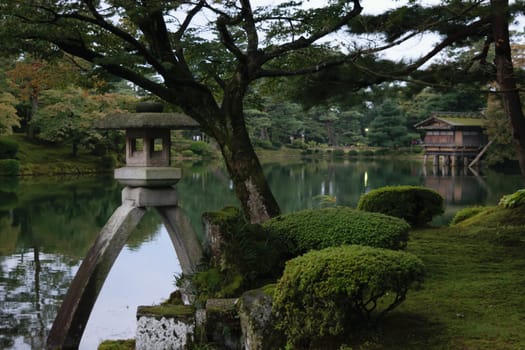 A serene Japanese garden with a stone lantern, lush greenery, and a calm pond reflecting the surrounding trees. A traditional wooden building is visible in the background.