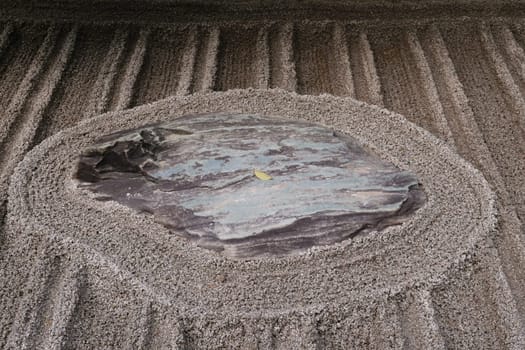 A close-up of a Zen garden with meticulously raked gravel patterns surrounding a central stone. The stone has a smooth, polished surface with natural striations.